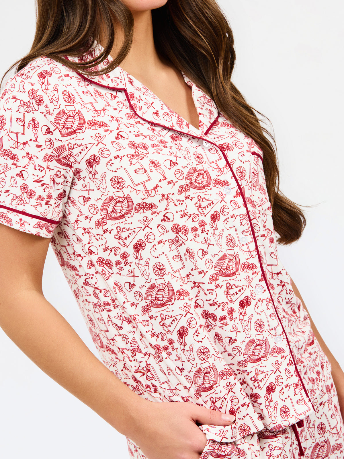 A woman modeling a two-piece pajama set with a short-sleeve button-front top and matching pants, featuring a pink and red floral pattern on a white background with dark red piping and a drawstring waist.