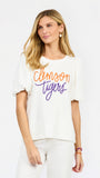 A woman modeling a white short-sleeve t-shirt with 'Clemson Tigers' printed across the chest in orange and purple script, paired with white pants against a light gray studio background.