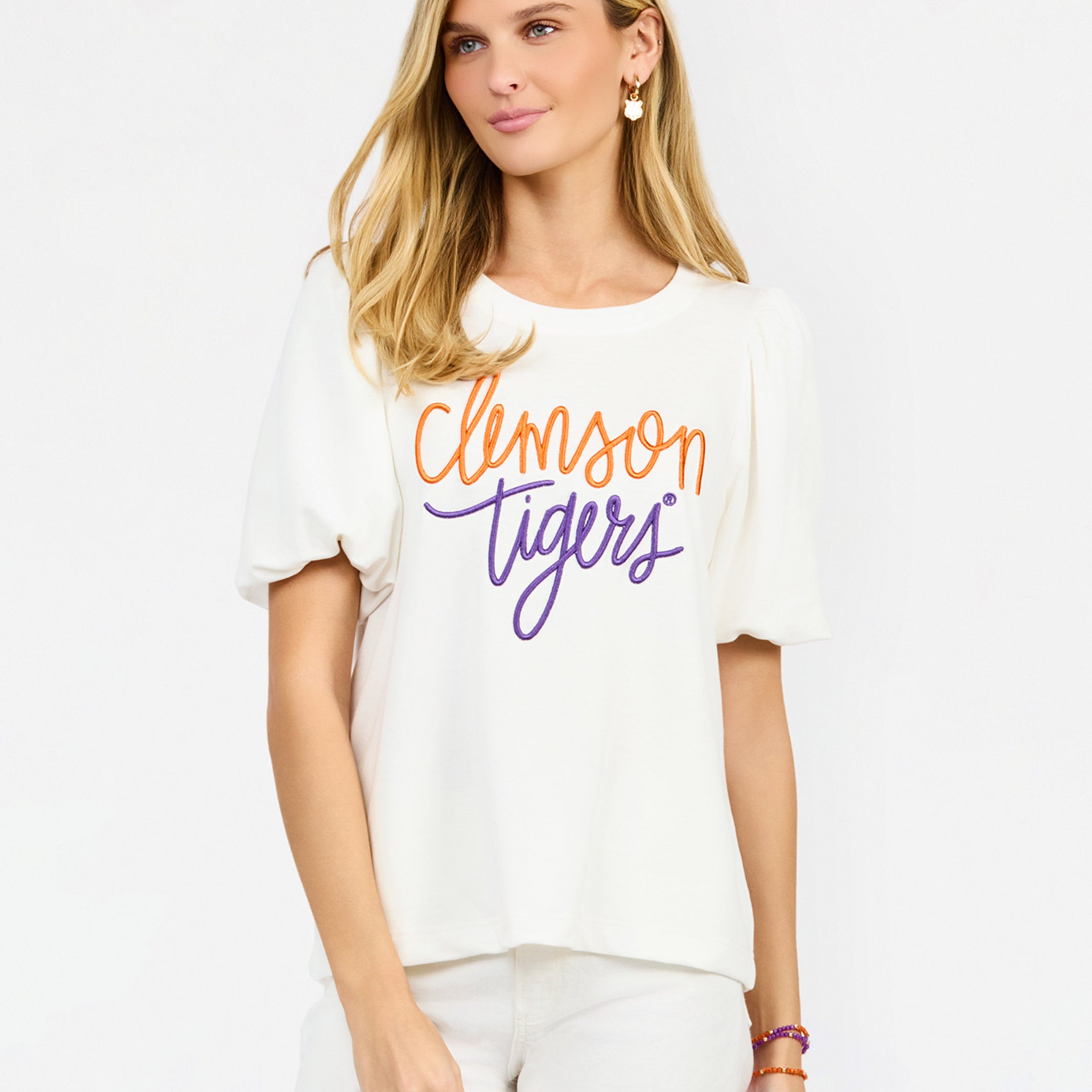 A woman modeling a white short-sleeve t-shirt with 'Clemson Tigers' printed across the chest in orange and purple script, paired with white pants against a light gray studio background.