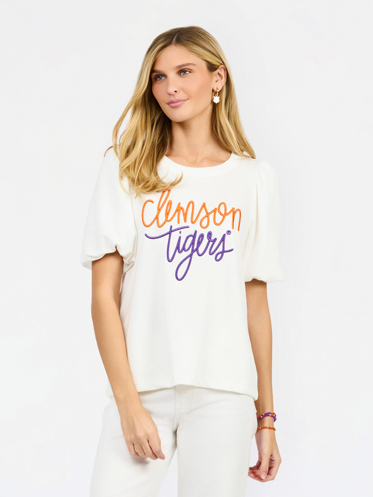 A woman modeling a white short-sleeve t-shirt with 'Clemson Tigers' printed across the chest in orange and purple script, paired with white pants against a light gray studio background.