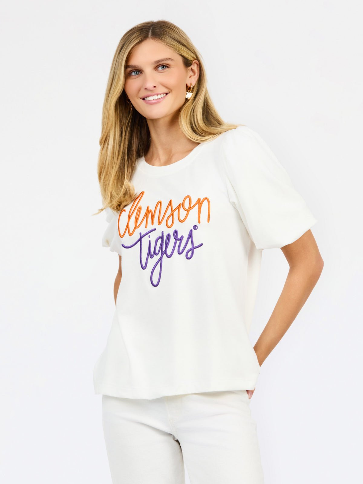 A woman wearing a white short-sleeve t-shirt with 'Clemson Tigers' printed in orange and purple script across the chest, paired with white pants, against a plain white background.