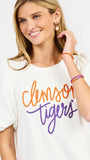 A woman wearing a white short-sleeve t-shirt with 'Clemson Tigers' printed in orange and purple script across the chest, paired with white pants, against a plain white background.
