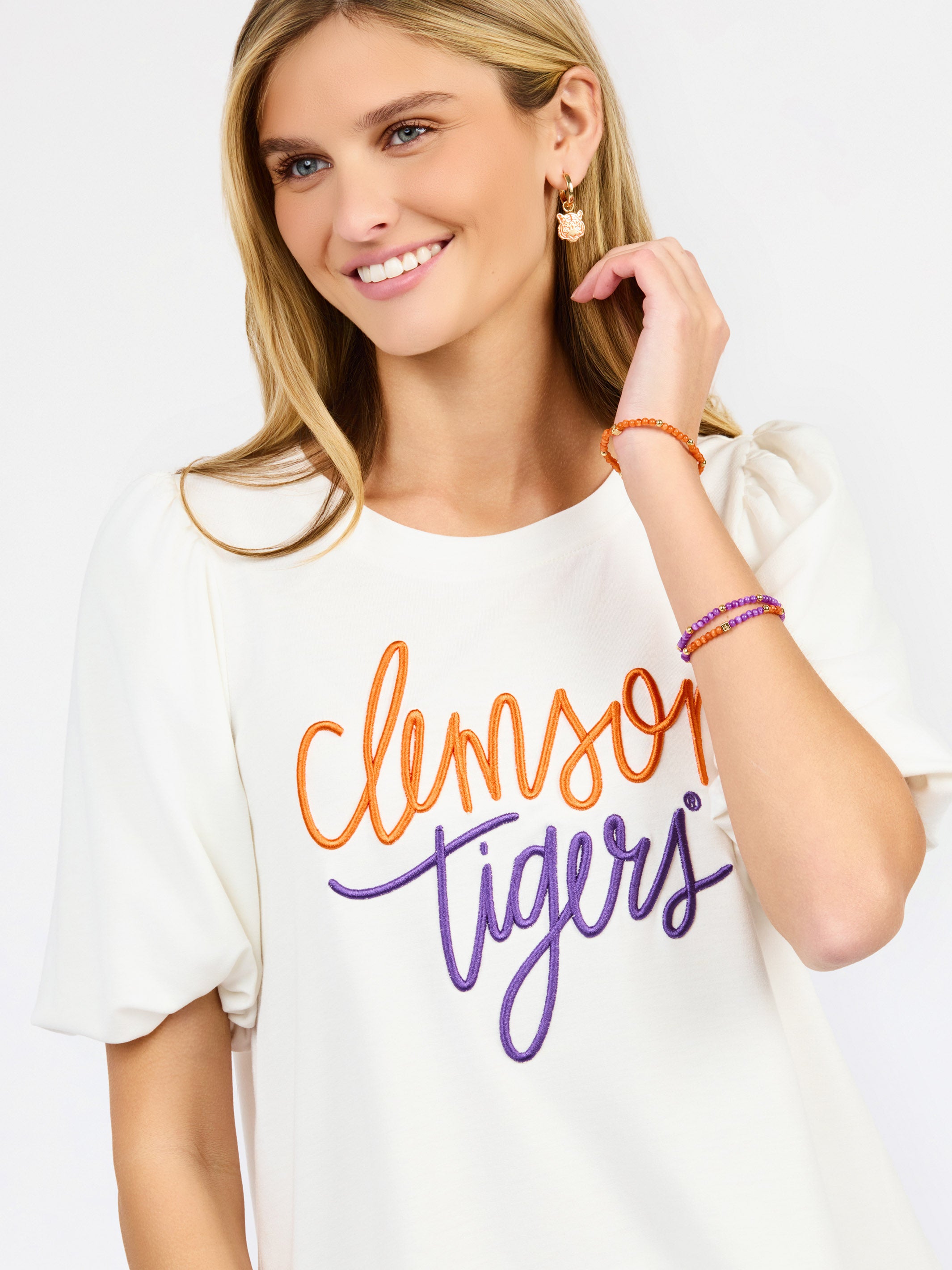 A woman wearing a white short-sleeve t-shirt with 'Clemson Tigers' printed in orange and purple script across the chest, paired with white pants, against a plain white background.