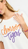 A woman wearing a white short-sleeve t-shirt with 'Clemson Tigers' printed in orange and purple script across the chest, paired with white pants, against a plain white background.