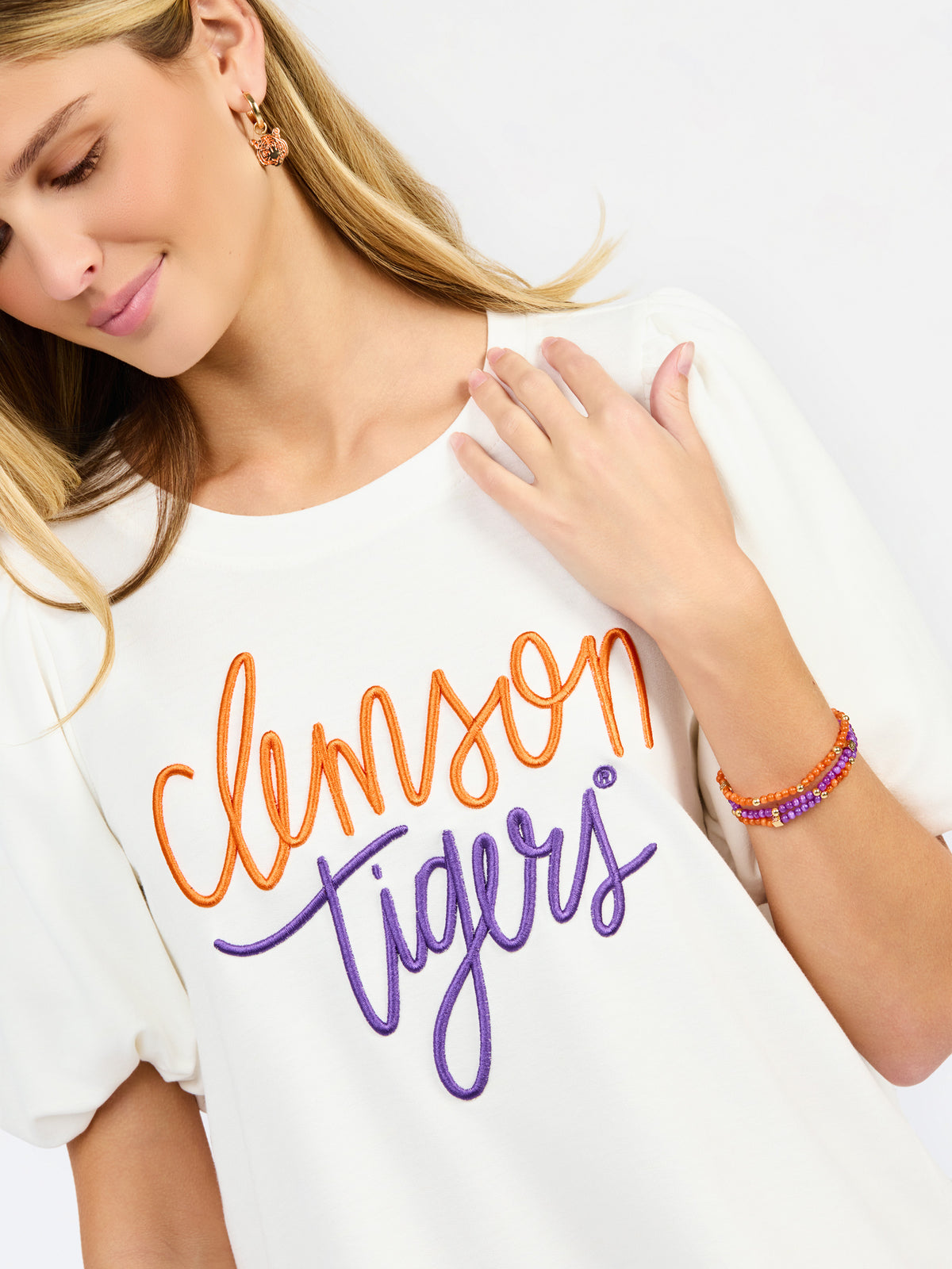 A woman wearing a white short-sleeve t-shirt with 'Clemson Tigers' printed in orange and purple script across the chest, paired with white pants, against a plain white background.