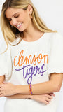 A woman wearing a white short-sleeve t-shirt with 'Clemson Tigers' printed in orange and purple script across the chest, paired with white pants, against a plain white background.