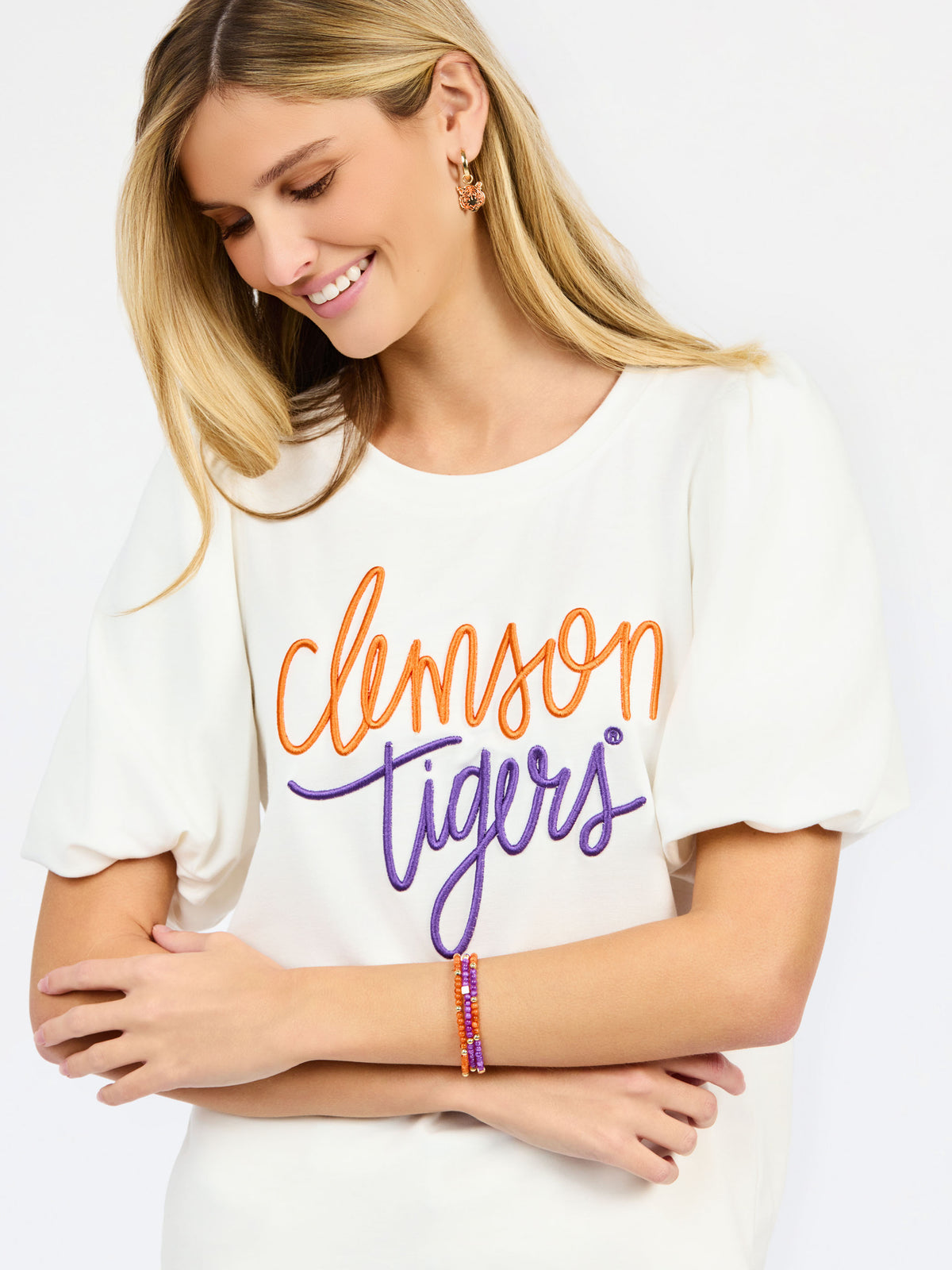 A woman wearing a white short-sleeve t-shirt with 'Clemson Tigers' printed in orange and purple script across the chest, paired with white pants, against a plain white background.
