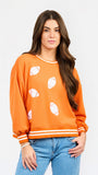 A woman modeling an orange crewneck sweatshirt with white football graphics and striped ribbed cuffs and hem, paired with blue jeans against a light gray background.