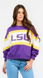 A woman wearing a purple, white, and yellow LSU sweatshirt with blue jeans, photographed against a plain light gray studio background.