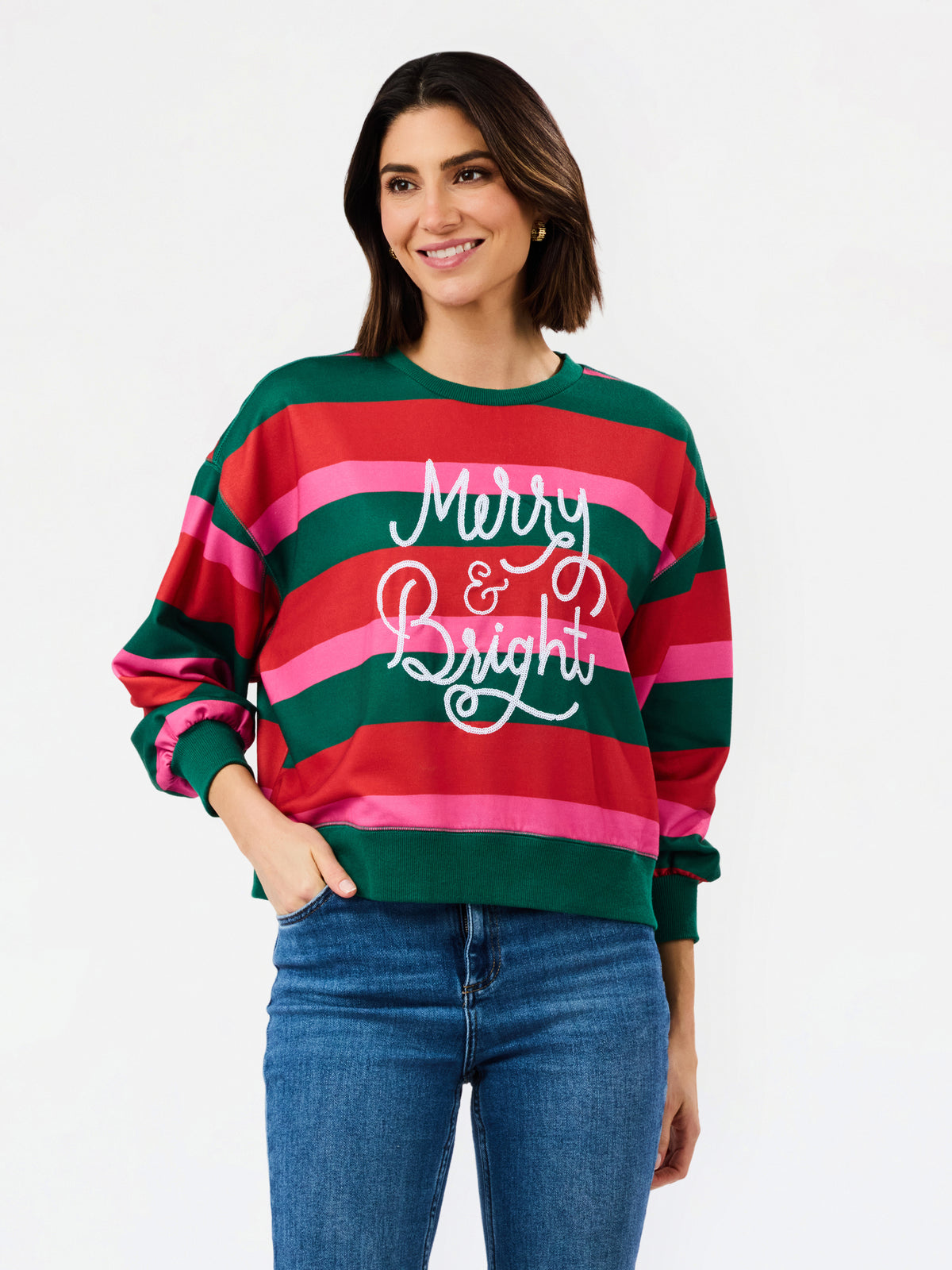 A woman wearing a festive holiday striped sweater with green, red, and pink horizontal stripes and 'Merry & Bright' written in white script across the chest, paired with blue jeans against a plain white background.