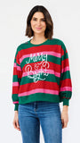 A woman wearing a festive holiday striped sweater with green, red, and pink horizontal stripes and 'Merry & Bright' written in white script, paired with blue jeans, against a plain white background.