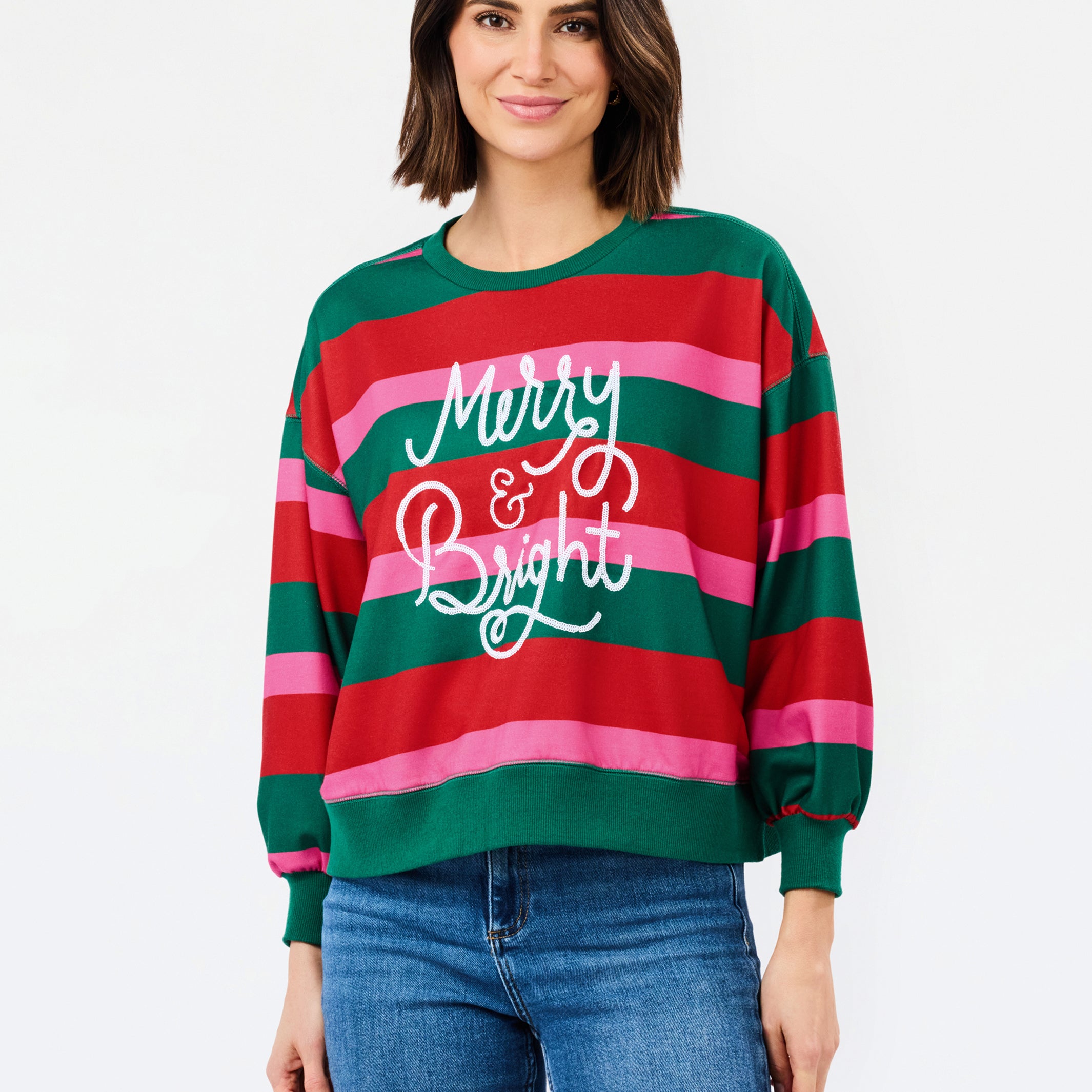 A woman wearing a festive holiday striped sweater with green, red, and pink horizontal stripes and 'Merry & Bright' written in white script, paired with blue jeans, against a plain white background.