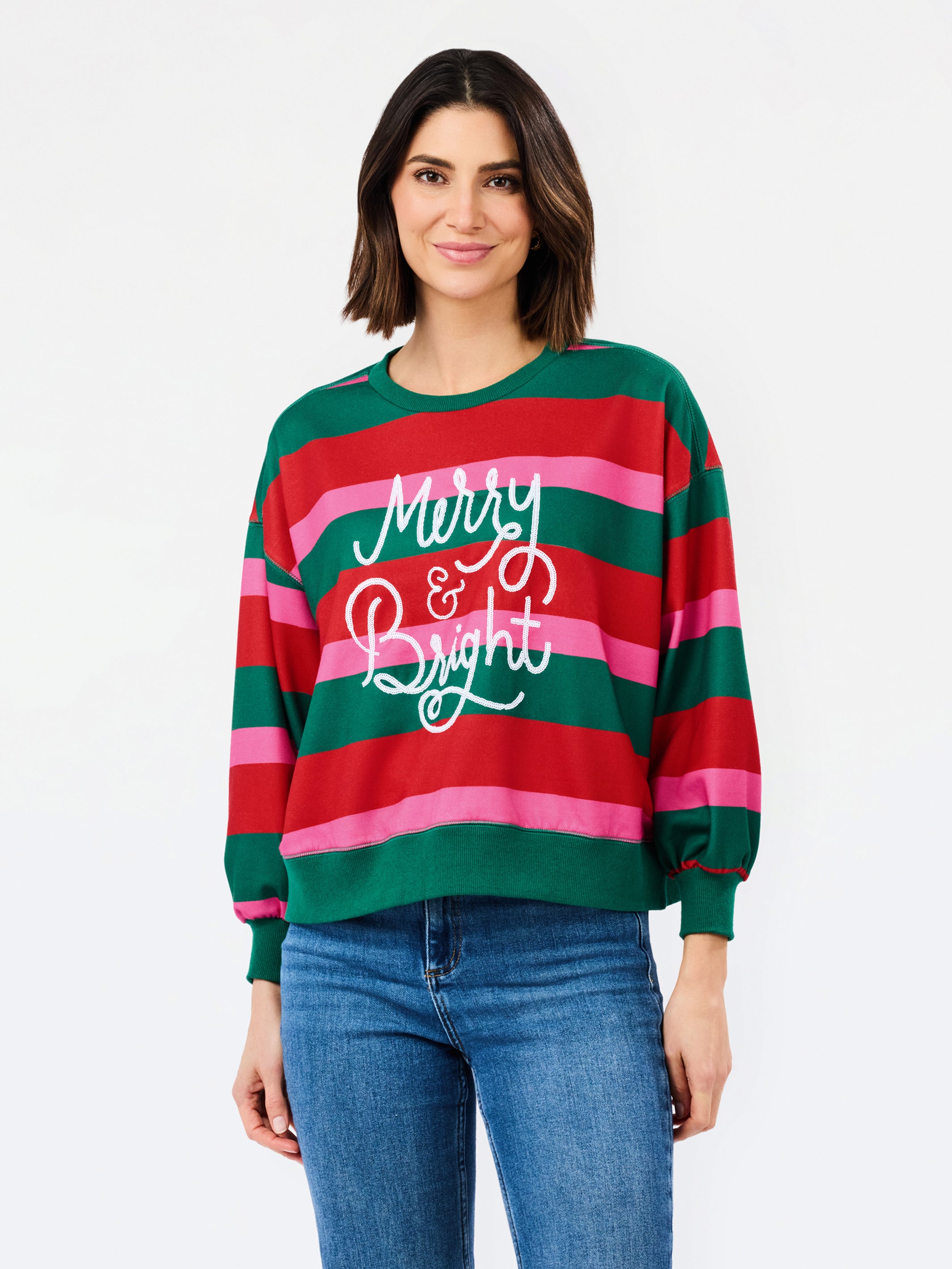 A woman wearing a festive holiday striped sweater with green, red, and pink horizontal stripes and 'Merry & Bright' written in white script, paired with blue jeans, against a plain white background.