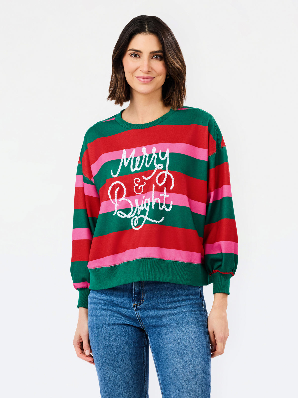 A woman wearing a festive holiday striped sweater with green, red, and pink horizontal stripes and 'Merry & Bright' written in white script, paired with blue jeans, against a plain white background.