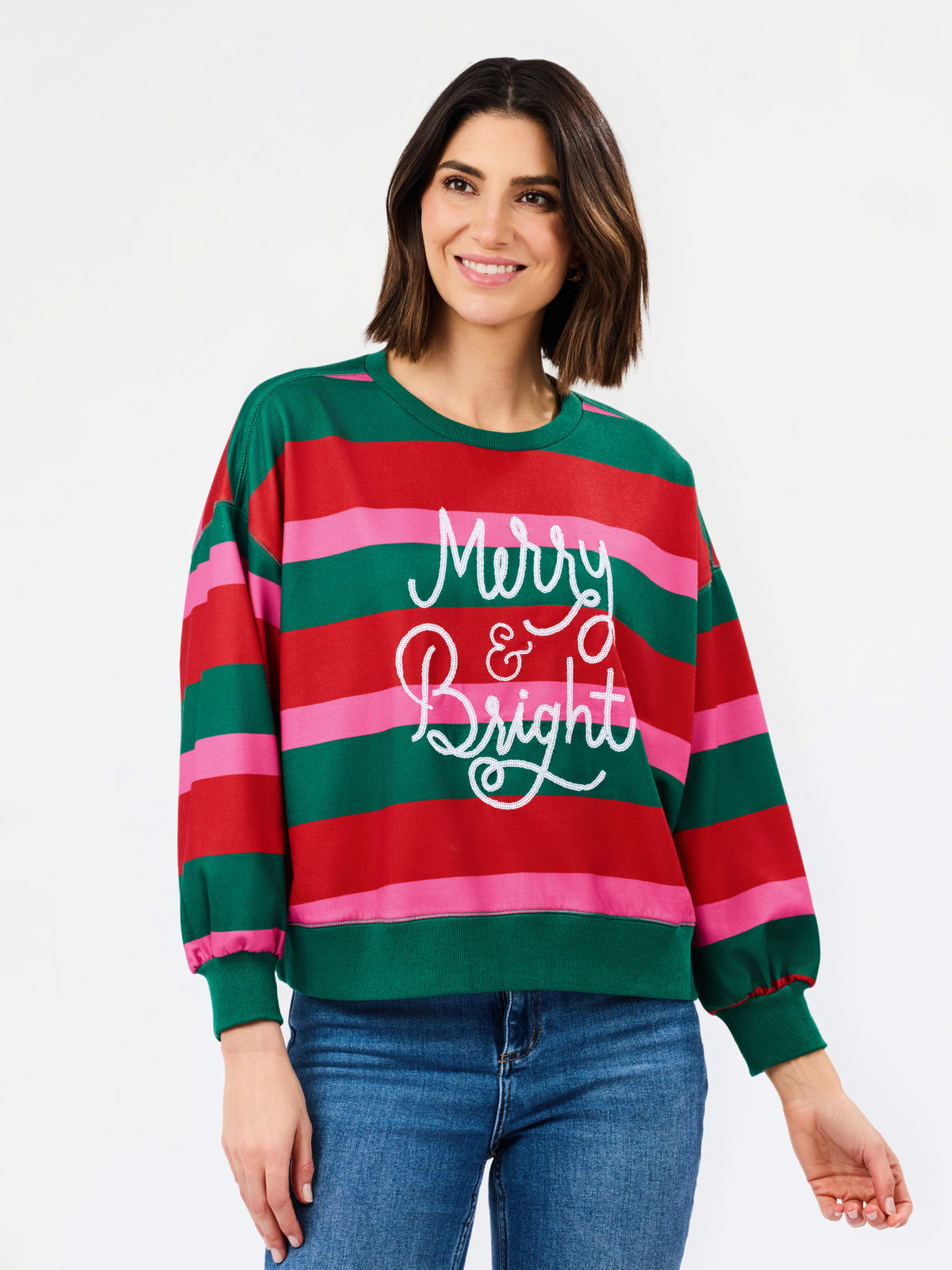 A woman wearing a festive crewneck sweatshirt with horizontal red, green, and pink stripes, featuring the text 'Merry & Bright' in white script across the chest. She is paired with blue jeans against a clean white background.