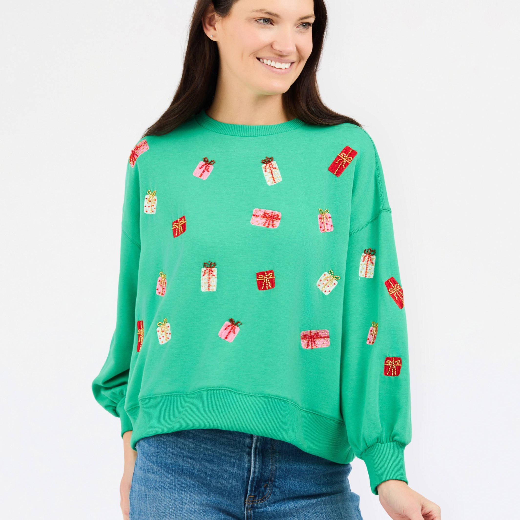 A woman wearing a bright green crewneck sweatshirt decorated with small red and white gift box motifs, paired with blue jeans, against a plain white studio background.