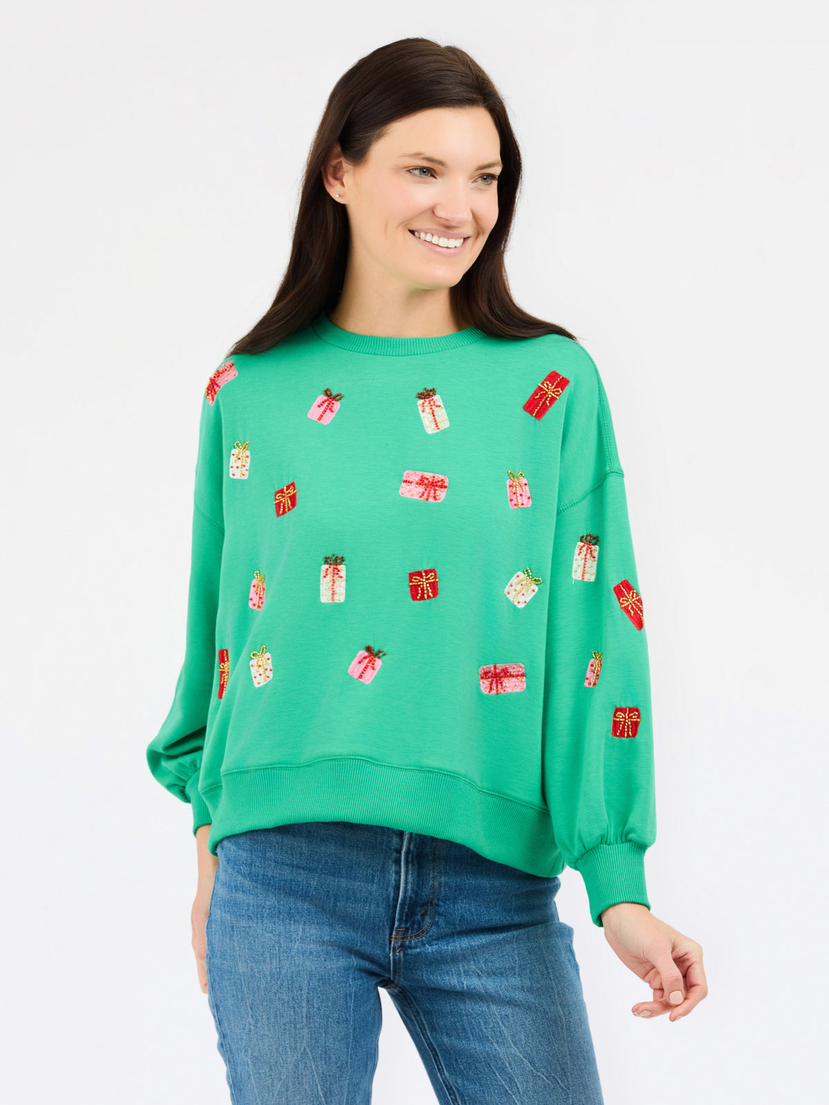 A woman wearing a bright green crewneck sweatshirt decorated with small red and white gift box motifs, paired with blue jeans, against a plain white studio background.