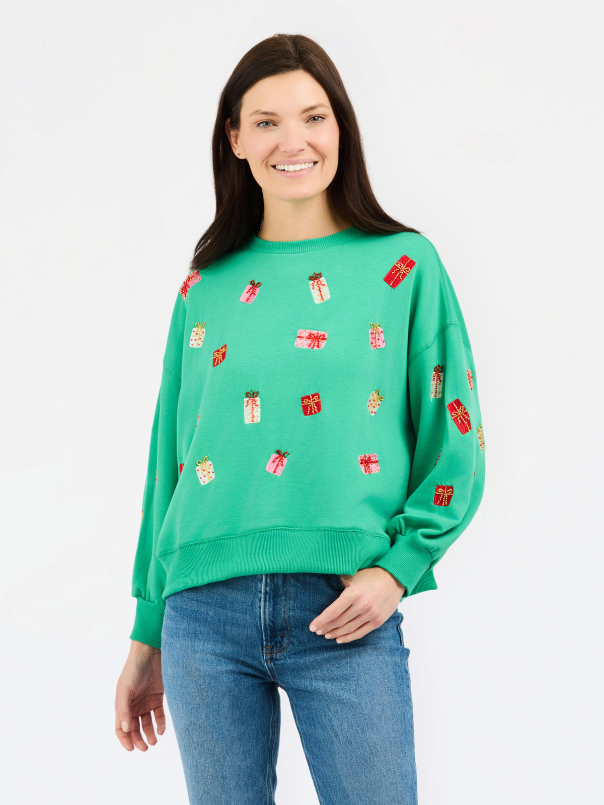 A woman wearing a bright green crewneck sweatshirt decorated with small red and gold gift box motifs, paired with blue jeans, against a plain white studio background.