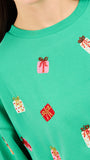 A close-up of a person wearing a bright green crewneck sweatshirt with an all-over festive pattern of embroidered gift boxes in red, pink, and white.
