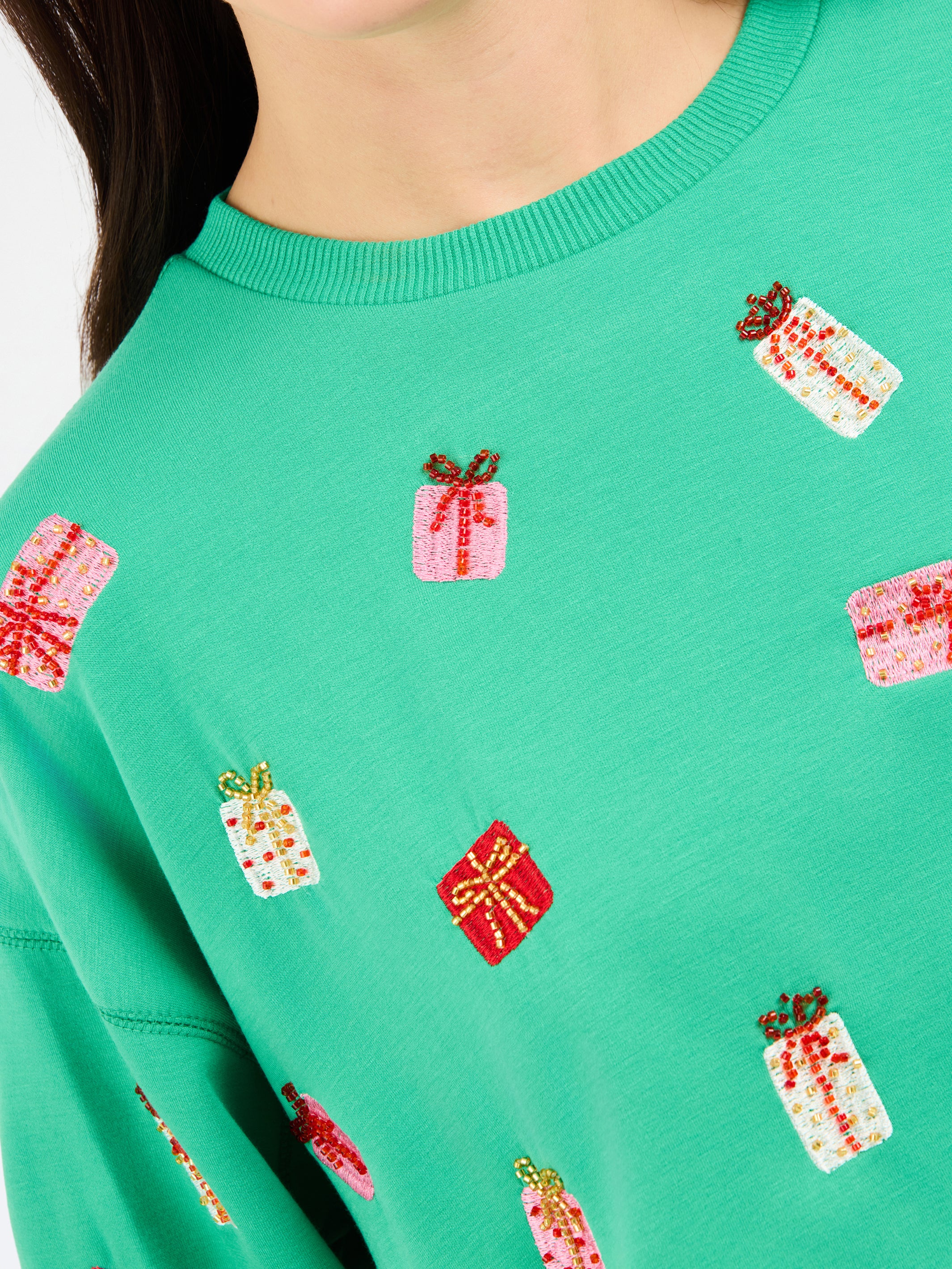A close-up of a person wearing a bright green crewneck sweatshirt with an all-over festive pattern of embroidered gift boxes in red, pink, and white.