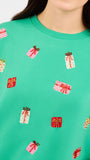 A close-up of a person wearing a bright green crewneck sweatshirt decorated with small sequined gift box motifs in pink, red, and white.