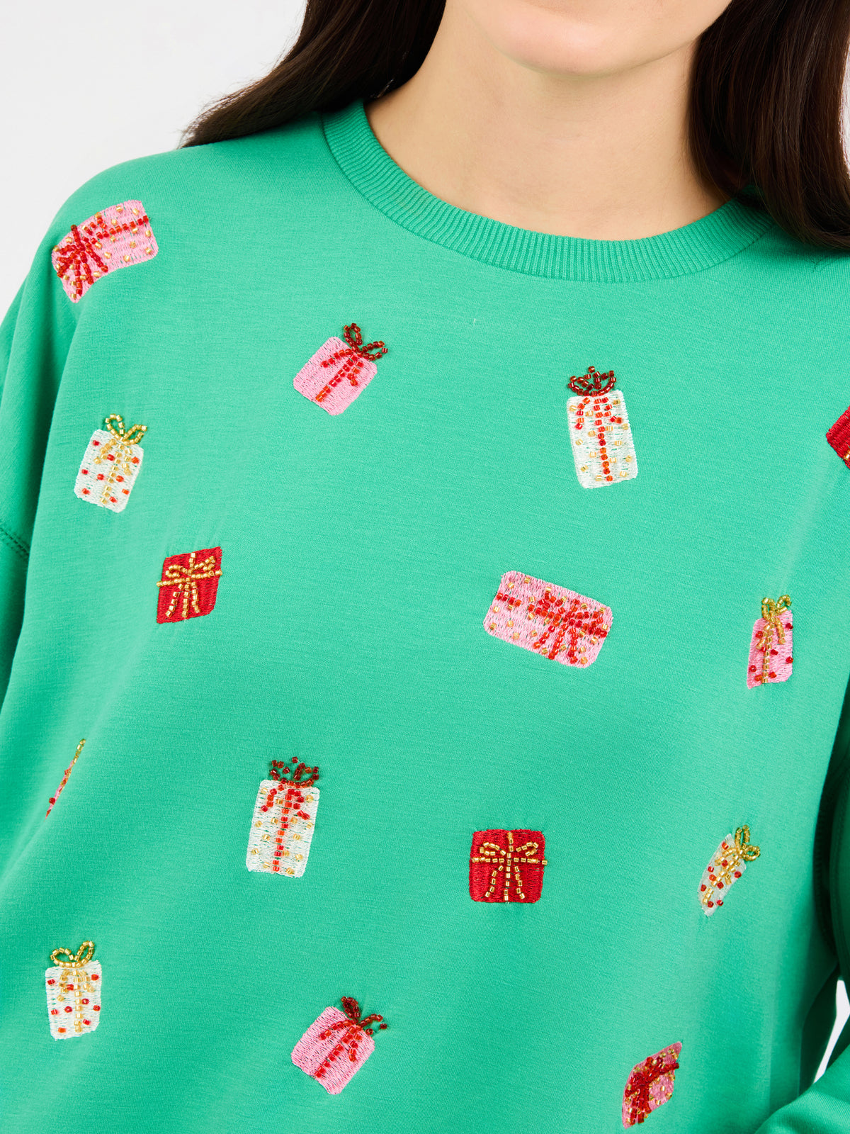 A close-up of a person wearing a bright green crewneck sweatshirt decorated with small sequined gift box motifs in pink, red, and white.