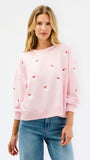 A woman wearing a light pink crewneck sweatshirt with small red and white embroidered santa motifs, paired with blue jeans, against a plain white studio background.