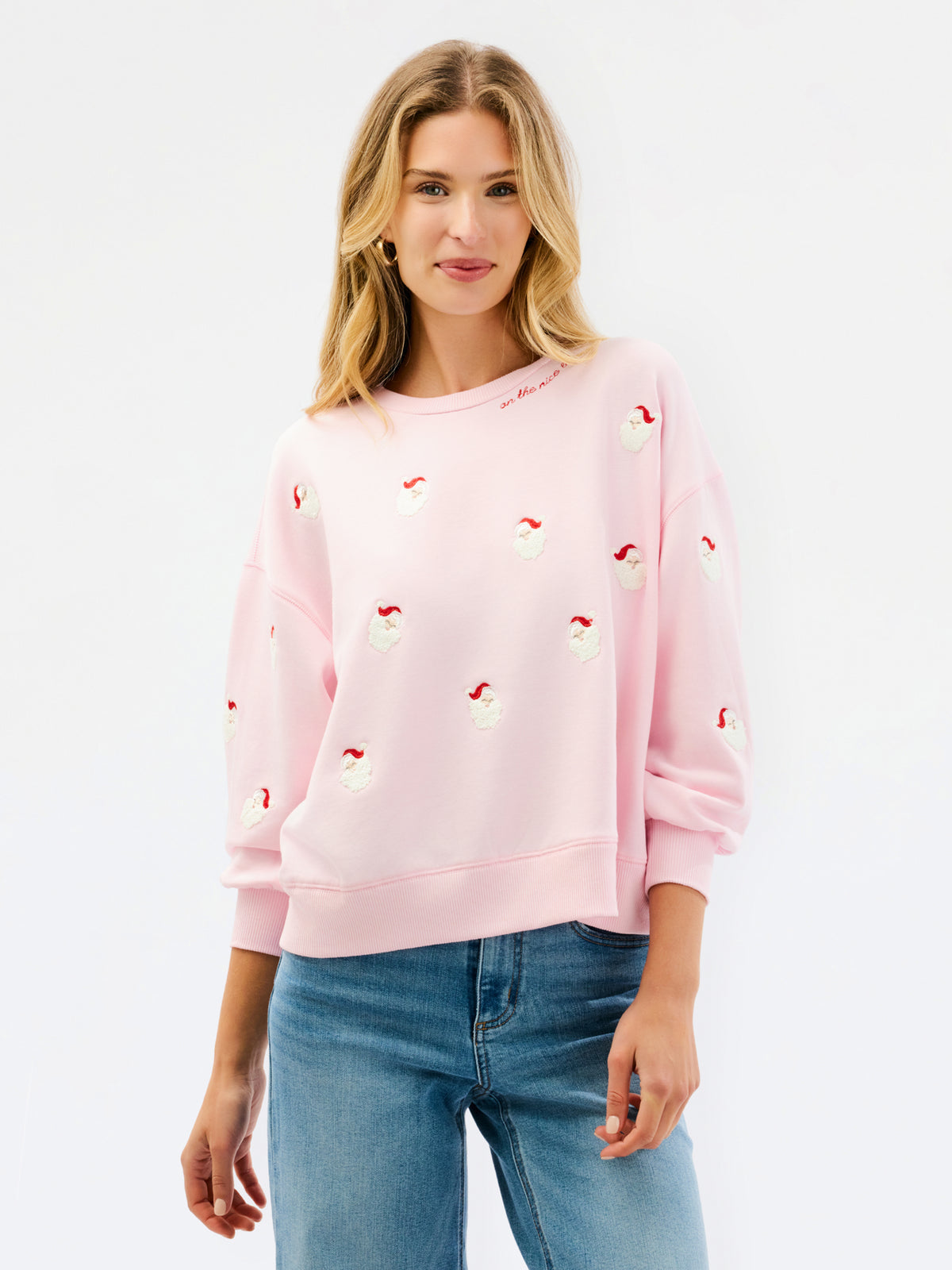 A woman wearing a light pink crewneck sweatshirt with small red and white embroidered santa motifs, paired with blue jeans, against a plain white studio background.
