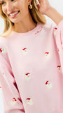 A woman wearing a light pink crewneck sweatshirt with small red and white embroidered santa motifs, paired with blue jeans, against a plain white studio background.