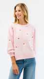 A woman wearing a light pink crewneck sweatshirt with small red and white embroidered santa motifs, paired with blue jeans, against a plain white studio background.