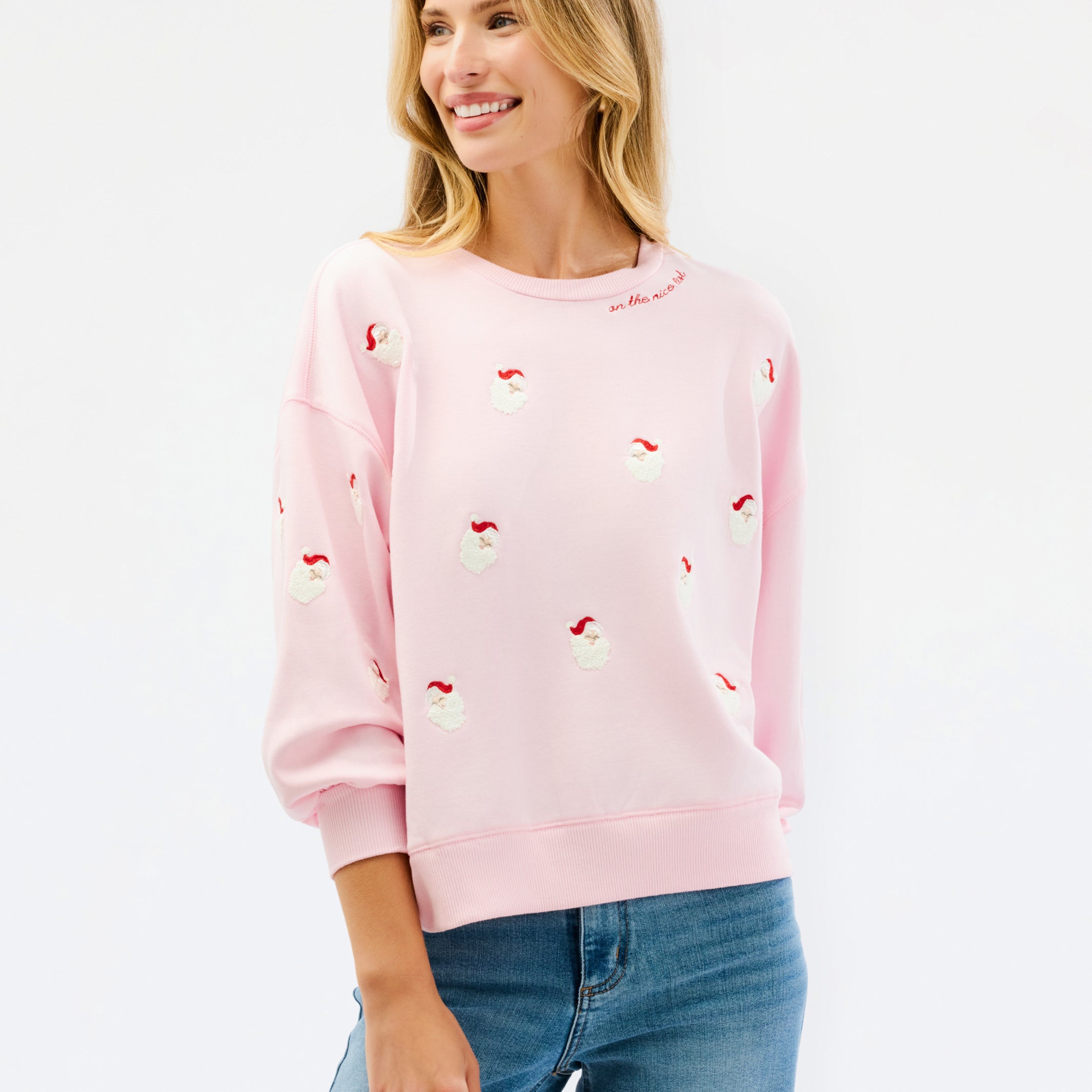 A woman wearing a light pink crewneck sweatshirt with small red and white embroidered santa motifs, paired with blue jeans, against a plain white studio background.