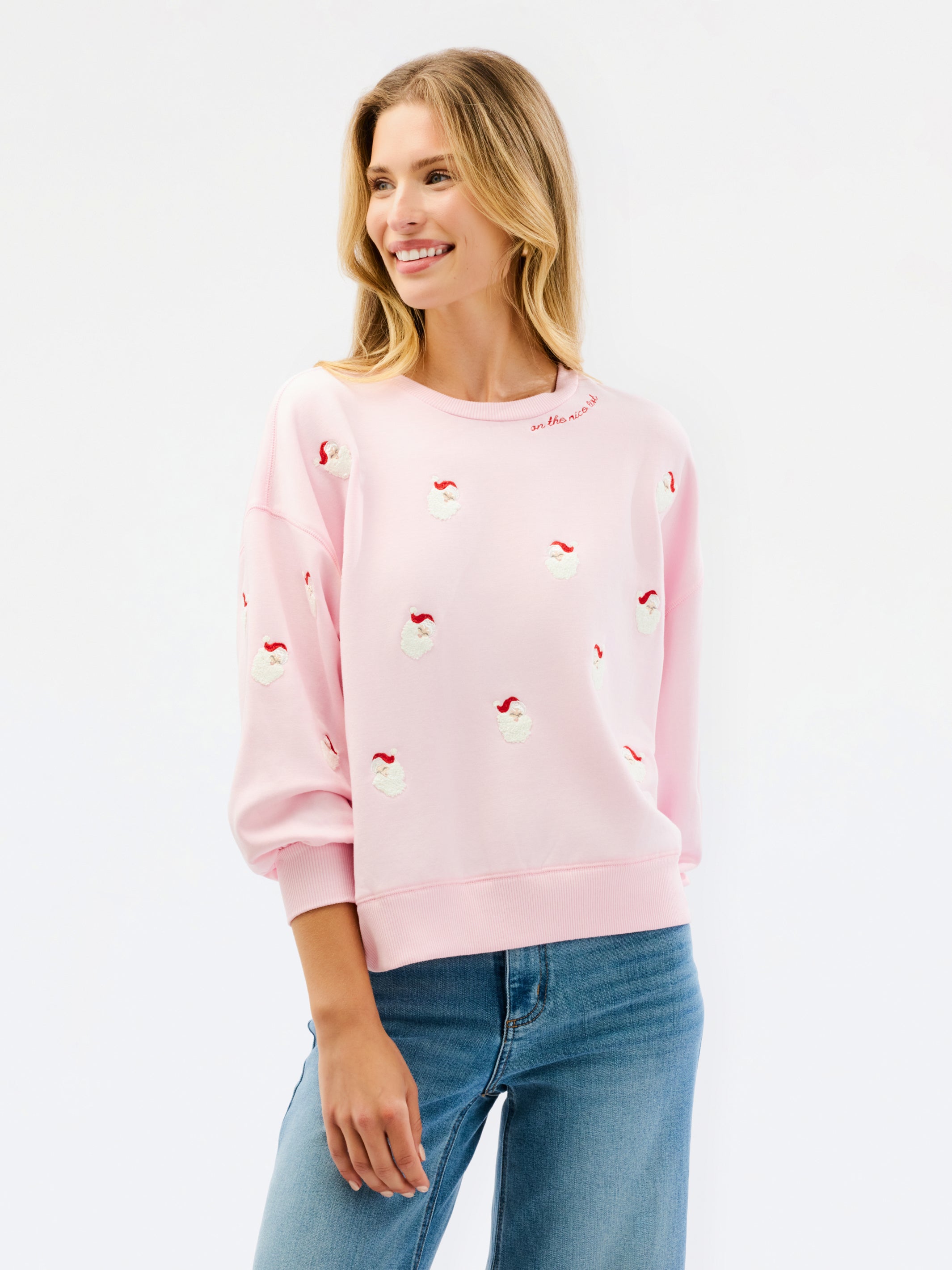 A woman wearing a light pink crewneck sweatshirt with small red and white embroidered santa motifs, paired with blue jeans, against a plain white studio background.