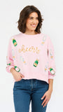 A woman wearing a light pink crewneck sweatshirt with 'cheers' written in gold script and decorated with champagne bottle graphics, paired with blue jeans, against a plain white background.