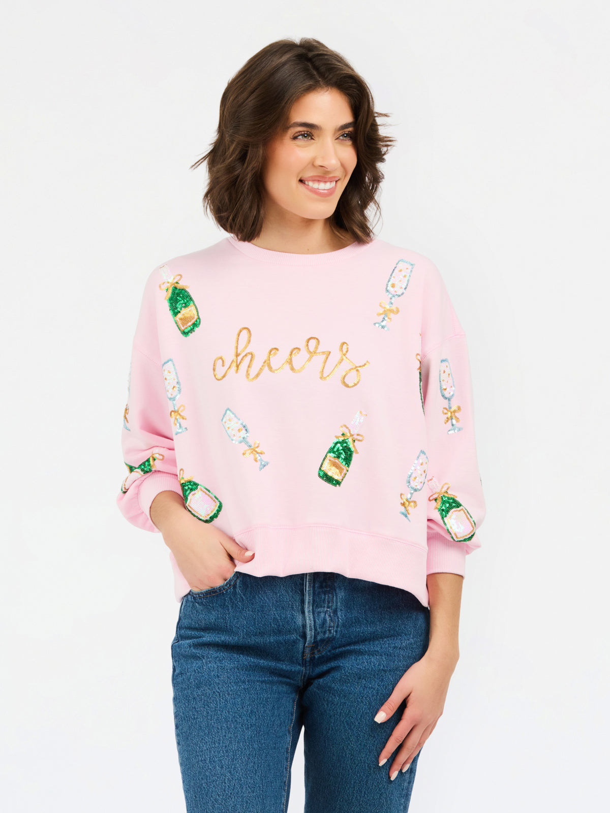 A woman wearing a light pink crewneck sweatshirt with 'cheers' written in gold script and decorated with champagne bottle graphics, paired with blue jeans, against a plain white background.