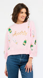 A woman wearing a light pink crewneck sweatshirt with 'cheers' embroidered in gold across the chest and green champagne bottle graphics, paired with blue jeans, against a plain white background.