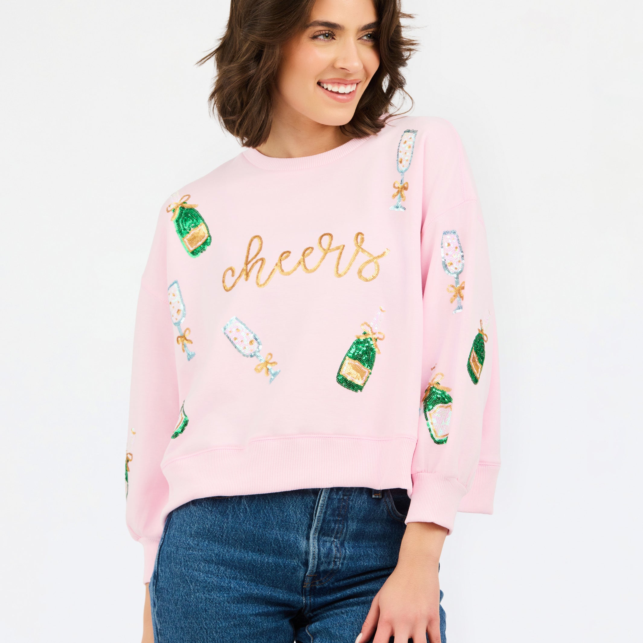 A woman wearing a light pink crewneck sweatshirt with 'cheers' embroidered in gold across the chest and green champagne bottle graphics, paired with blue jeans, against a plain white background.