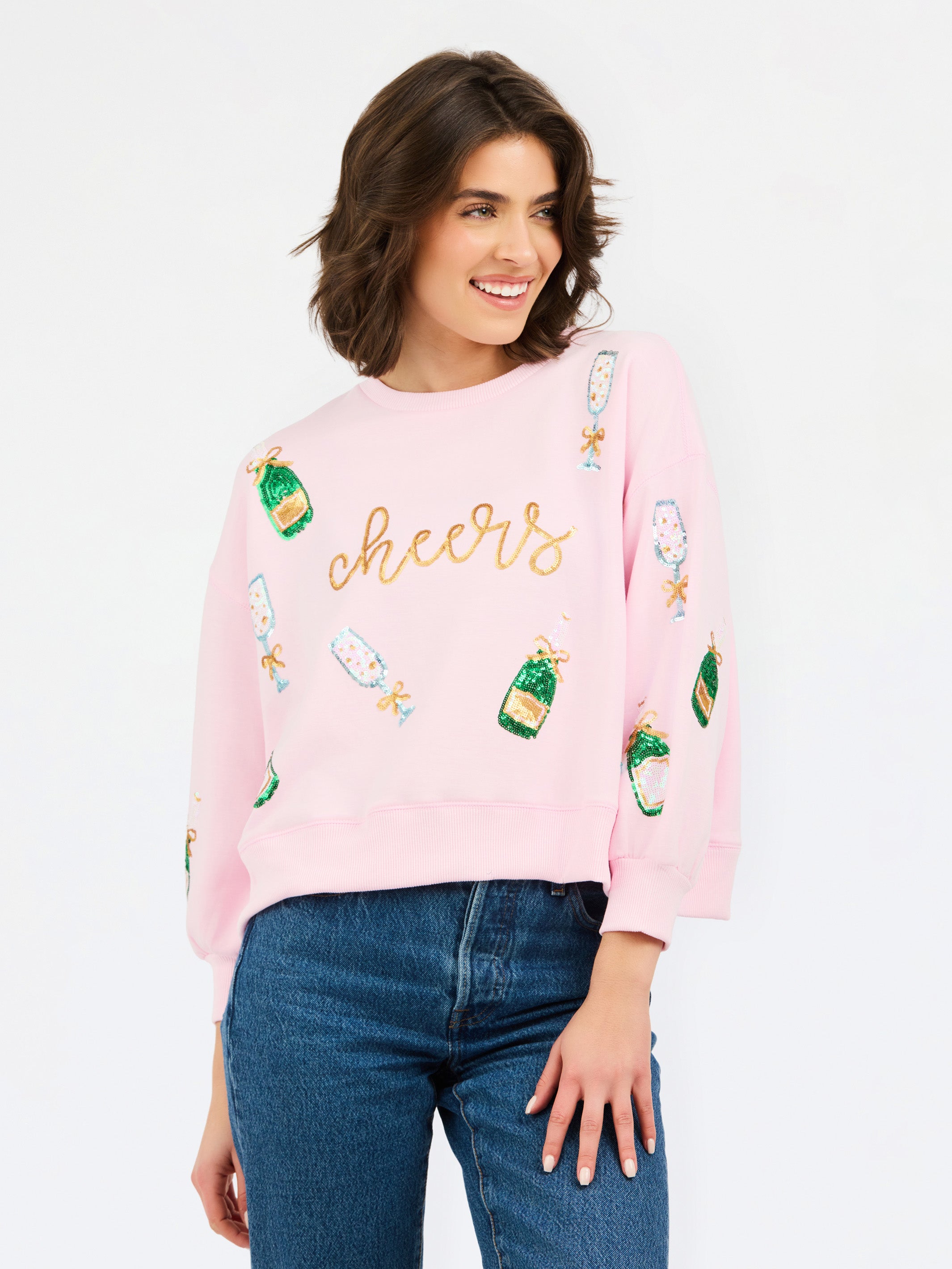 A woman wearing a light pink crewneck sweatshirt with 'cheers' embroidered in gold across the chest and green champagne bottle graphics, paired with blue jeans, against a plain white background.