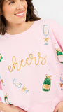 A woman wearing a light pink crewneck sweatshirt with 'cheers' written in gold glitter across the chest, decorated with sequined champagne bottles and glasses.