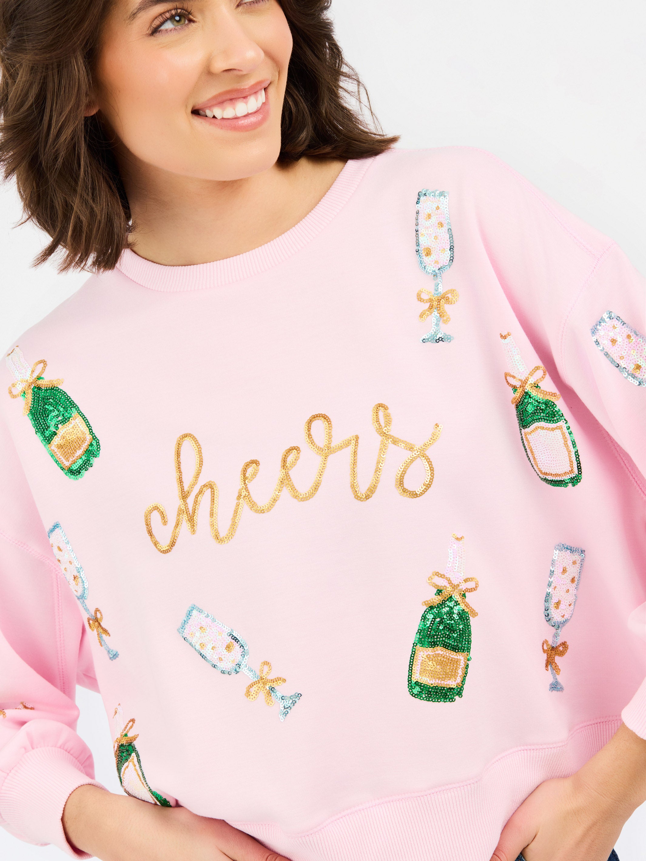 A woman wearing a light pink crewneck sweatshirt with 'cheers' written in gold glitter across the chest, decorated with sequined champagne bottles and glasses.