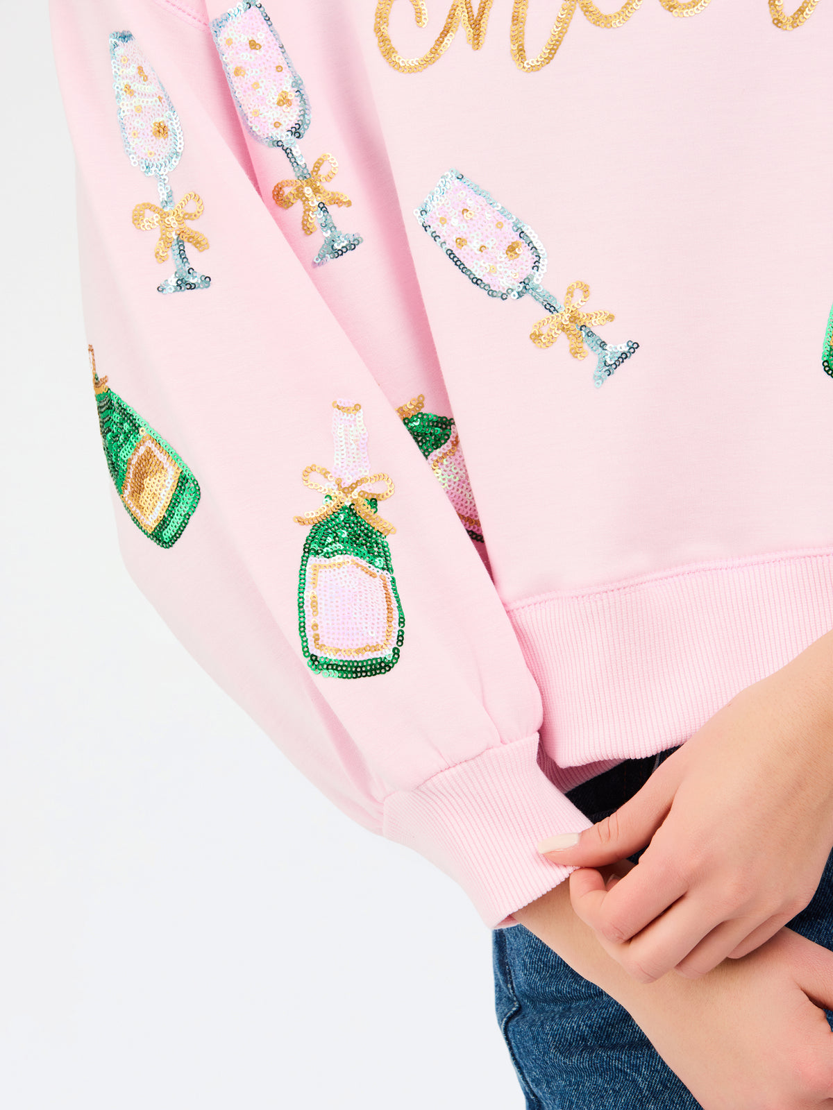 A close-up of a light pink sweatshirt featuring sequined champagne bottle and glass motifs on the sleeves, worn by a model whose hands and jeans are partially visible.