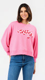 A woman wearing a pink crewneck sweatshirt with red script embroidery spelling 'merry' on the front, paired with blue jeans, against a plain white studio background.
