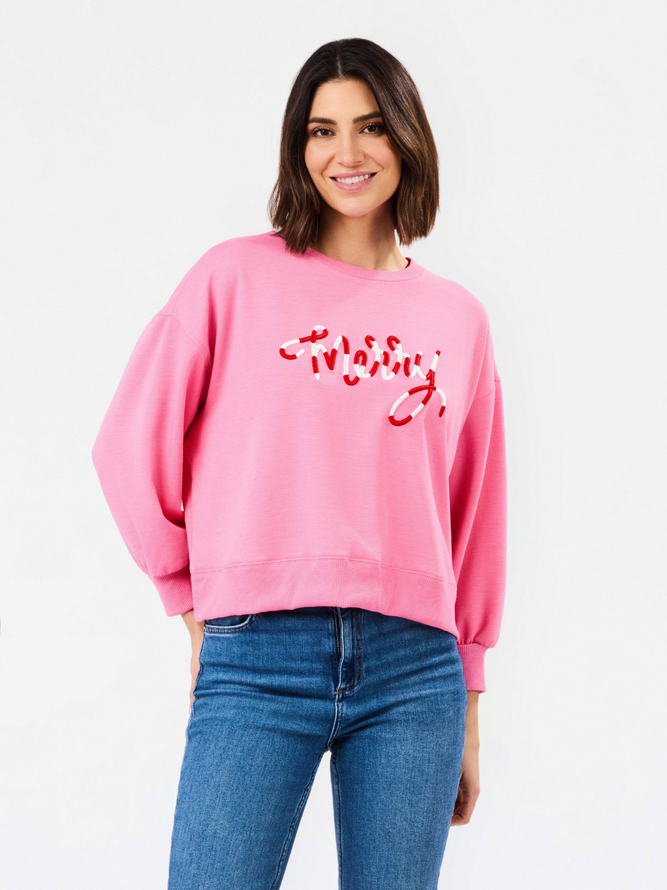 A woman wearing a pink crewneck sweatshirt with red script embroidery spelling 'merry' on the front, paired with blue jeans, against a plain white studio background.