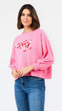 A woman wearing a pink crewneck sweatshirt with red script embroidery spelling 'merry' on the front, paired with blue jeans, against a plain white studio background.