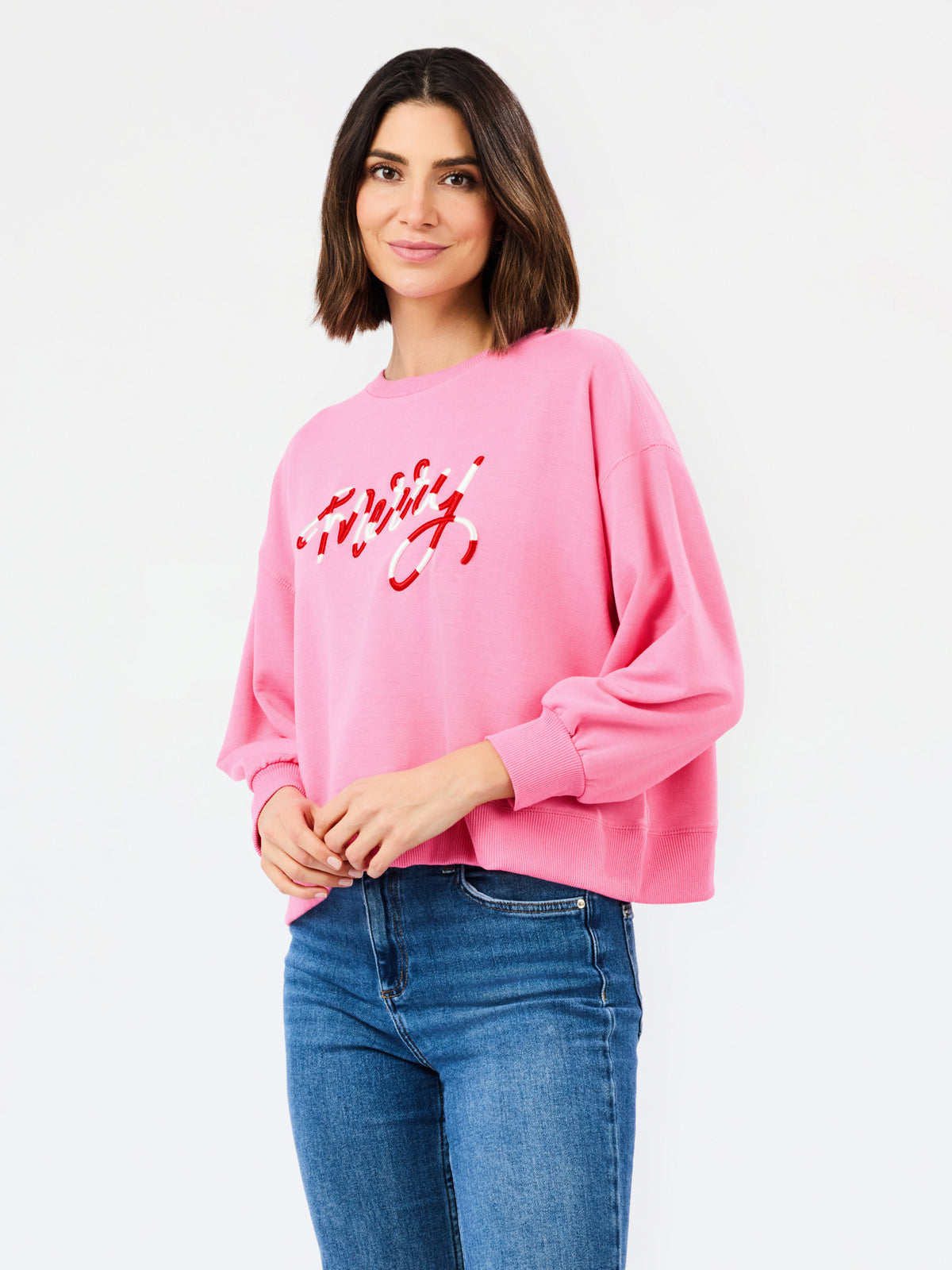 A woman wearing a pink crewneck sweatshirt with red script embroidery spelling 'merry' on the front, paired with blue jeans, against a plain white studio background.
