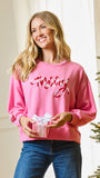 A woman wearing a pink crewneck sweatshirt with 'Merry' in red sequins, holding a small pink gift box with a white bow. She is standing indoors next to a decorated Christmas tree.