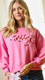 A woman wearing a pink crewneck sweatshirt with 'Merry' in red and white cursive embroidery, paired with dark blue jeans, holding a white gift bow in a holiday-themed indoor setting.