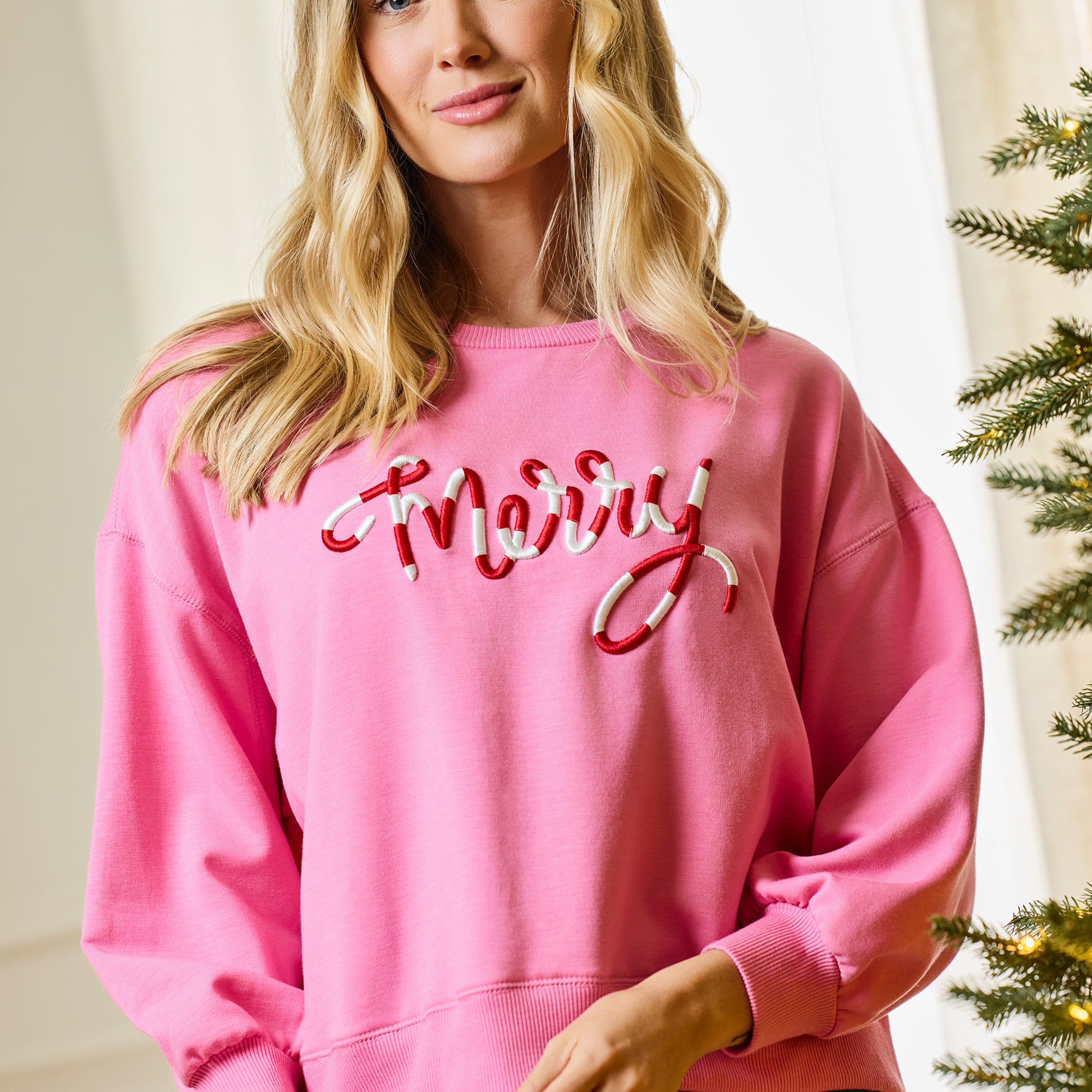 A woman wearing a pink crewneck sweatshirt with 'Merry' in red and white cursive embroidery, paired with dark blue jeans, holding a white gift bow in a holiday-themed indoor setting.