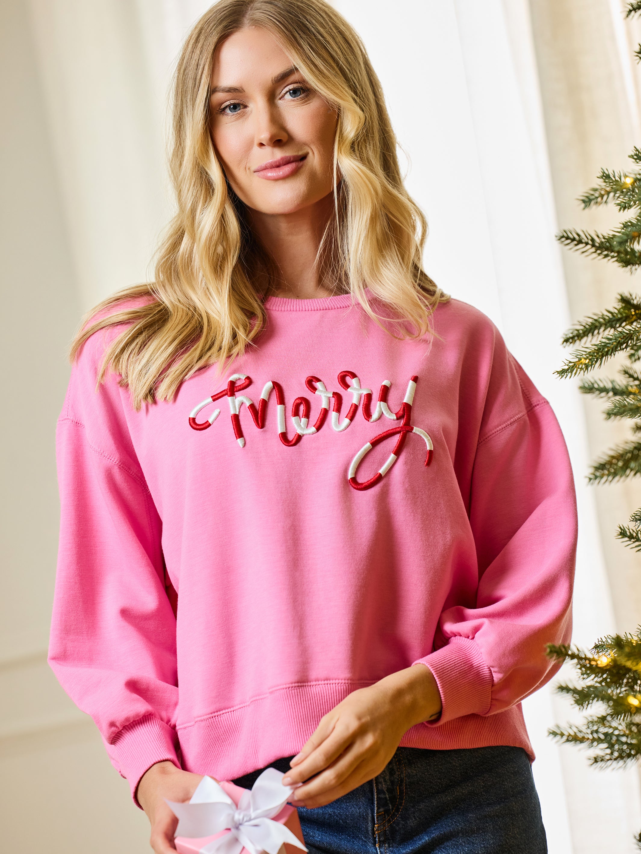A woman wearing a pink crewneck sweatshirt with 'Merry' in red and white cursive embroidery, paired with dark blue jeans, holding a white gift bow in a holiday-themed indoor setting.