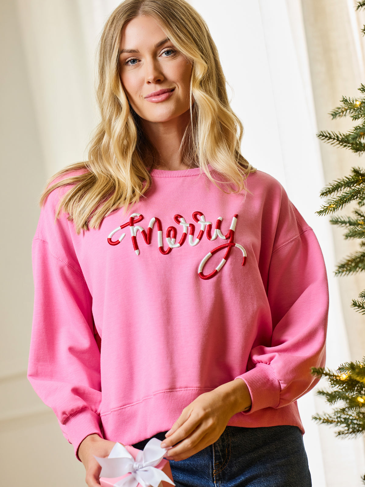 A woman wearing a pink crewneck sweatshirt with 'Merry' in red and white cursive embroidery, paired with dark blue jeans, holding a white gift bow in a holiday-themed indoor setting.