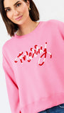 A woman wearing a pink crewneck sweatshirt with red script embroidery spelling 'merry' on the front, paired with blue jeans, against a plain white studio background.